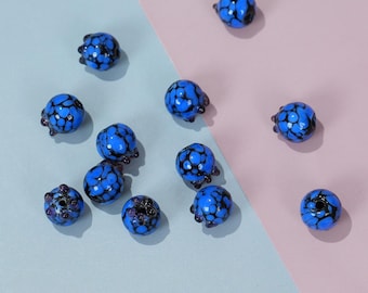 May include: A collection of blue glass beads with a speckled black pattern. Each bead has a rounded shape with small, dark protrusions. The beads are scattered on a two-tone background of light blue and pink.