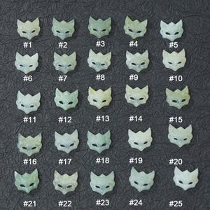 May include: Twenty-five pale green fox mask pendants, each numbered from #1 to #25, arranged in a grid on a dark gray surface. The masks have pointed ears and eye holes, suitable for jewelry making or crafts.