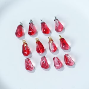 Könnte beinhalten: Eine Sammlung roter, tropfenförmiger Anhänger mit silber- und goldfarbenen Metallschlaufen. Die Anhänger sind durchscheinend und zeigen ein helleres rotes Innere. Die Anhänger sind auf einer weißen Oberfläche angeordnet.