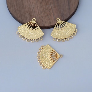 May include: Three gold-coloured fan-shaped jewellery findings. Each fan has a detailed floral design and multiple small loops along the bottom edge for attaching beads or other components. The fans are arranged on a grey surface.