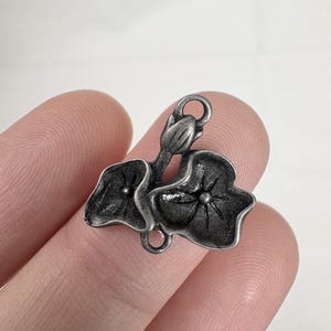 May include: A small, antique silver-toned metal charm featuring a floral design. The charm depicts two leaves and a bud, with a loop at the top for attaching to jewelry or other crafts. The leaves have detailed veining.