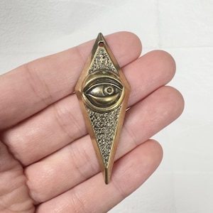 Könnte beinhalten: Ein rautenförmiger Anhänger mit Messing-Finish. Die Mitte zeigt ein detailliertes Augendesign, umgeben von einem strukturierten Rand. Der Anhänger hat oben ein kleines Loch zum Aufhängen.