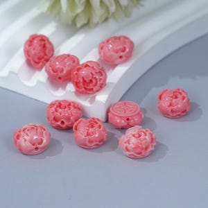 Peut inclure: Des perles roses en forme de pivoine avec des détails floraux complexes. Ces perles sont faites d'un matériau lisse et brillant et sont disposées sur une surface blanche sur un fond gris clair. Les perles sont d'une teinte rose uniforme.