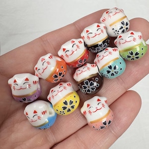 Può includere: Una collezione di piccole statuette Maneki-neko in ceramica. Ogni gatto ha un viso bianco con dettagli rossi, un colletto dorato e una base colorata decorata con motivi floreali. Le basi sono in vari colori, tra cui arancione, giallo e blu.