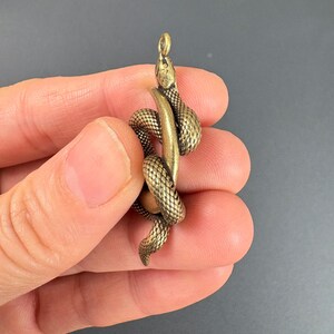 May include: A detailed brass snake pendant coiled around a cylindrical form. The snake has a textured, scale-like pattern and a small loop at the top for hanging. The pendant is a warm, antique gold color.