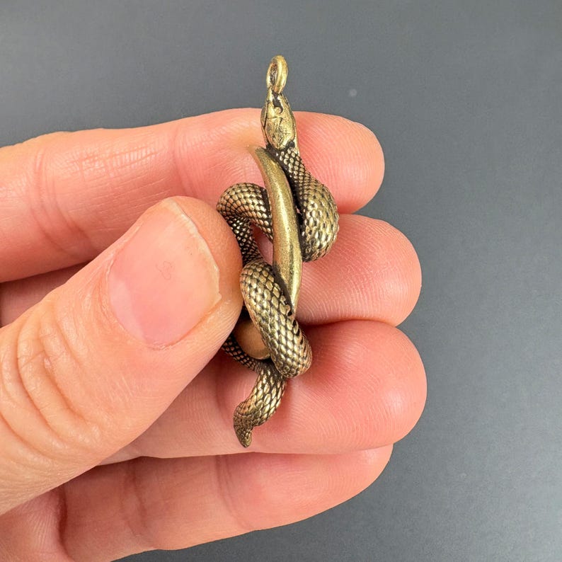 May include: A detailed brass snake pendant coiled around a cylindrical form. The snake has a textured, scale-like pattern and a small loop at the top for hanging. The pendant is a warm, antique gold color.
