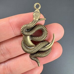 May include: Bronze-colored snake pendant with a coiled body and detailed scales. The pendant has a loop at the top for attaching to a chain or other jewelry. The snake's head is raised, and its tail curls upward.