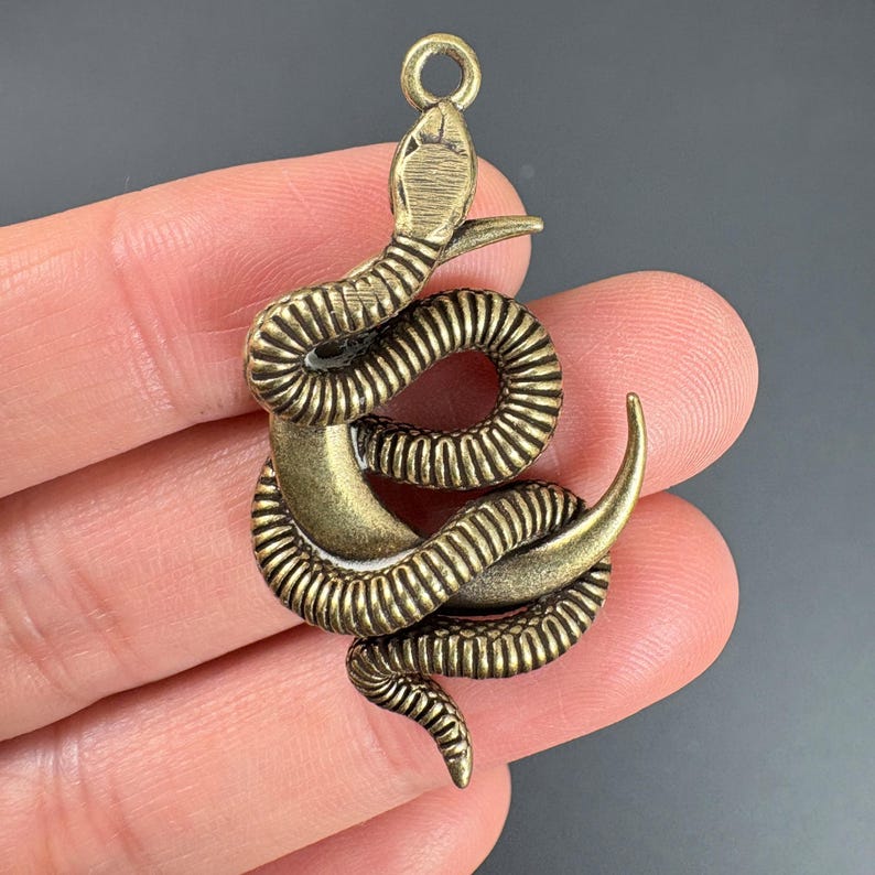 May include: Bronze-colored snake pendant with a coiled body and detailed scales. The pendant has a loop at the top for attaching to a chain or other jewelry. The snake's head is raised, and its tail curls upward.