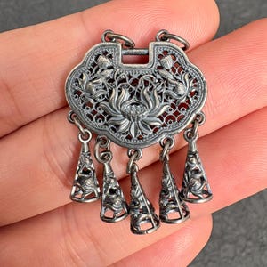 Op de afbeelding: Een zilverkleurige metalen hanger in de vorm van een traditioneel slot, voorzien van ingewikkeld opengewerkt filigraan en reliëfmotieven van lotusbloemen. Het sieraad heeft vijf kegelvormige bedels die met kleine ringetjes aan de basis hangen.