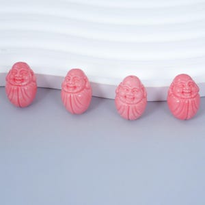 Puede incluir: Cuatro figuras rosas de Buda con caras sonrientes. Las figuras ovaladas tienen rasgos faciales y túnicas detalladas. El fondo es una superficie gris claro con una estructura blanca curvada.