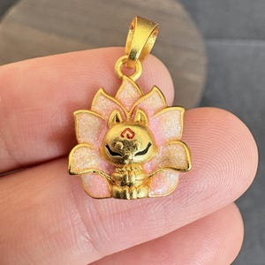 May include: A gold pendant with a cat design set within a lotus flower. The cat has black eyes and a red heart on its forehead. The lotus petals are pink with gold accents. The pendant has a loop for a chain.