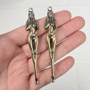 Könnte beinhalten: Zwei bronzefarbene Anhänger in Form einer nackten Frau. Jeder Anhänger hat oben eine Schlaufe zum Aufhängen und einen schlanken, länglichen Körper. Das Haar der Frau ist lang und wellig, und ihre Arme sind über der Brust verschränkt.
