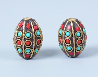 May include: Two decorative, oval-shaped beads. The beads are dark brown with gold-colored metal accents. They feature red and turquoise-colored stones in a repeating pattern. The beads have a hole at the top for stringing.