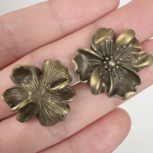 May include: Two antique brass-colored flower-shaped embellishments. Each flower has five petals with detailed veining and a central stamen. One flower has a loop on the back. The embellishments are likely for crafts or jewelry making.