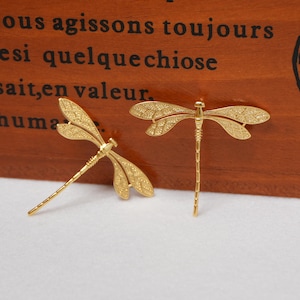 May include: Two gold-colored dragonfly brooches. The brooches are detailed with textured wings and slender bodies. The dragonfly brooches are displayed on a wooden surface with French text.