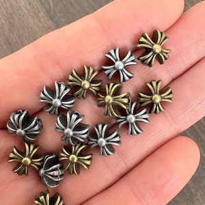 May include: A selection of small, metal cross-shaped beads in silver, gold, and bronze finishes. The beads feature an intricate, ornate design with multiple points radiating from a central sphere. The beads are displayed in a hand.