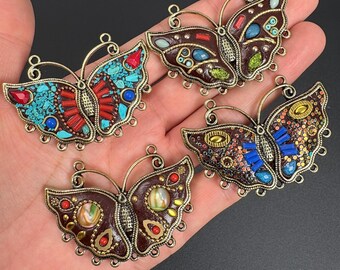 Könnte beinhalten: Vier dekorative Schmetterlingsanhänger mit aufwendigen Designs. Jeder Schmetterling hat einen bronzefarbenen Rahmen und Flügel, die mit farbigen Steinen und Mosaikmustern verziert sind. Die Flügel zeigen eine Vielzahl von Farben, darunter Türkis, Rot, Blau und Braun.