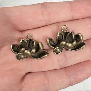 May include: Two bronze-colored lotus flower charms. Each charm has detailed petals and a small loop at the top for attaching to jewelry. The charms are designed to be used for crafting earrings, necklaces, or bracelets.