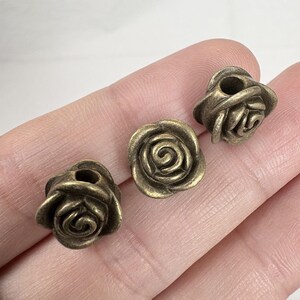 May include: Three bronze-colored rose-shaped beads. Each bead has detailed petals and a hole through the center. The beads are small and appear to be made of metal.