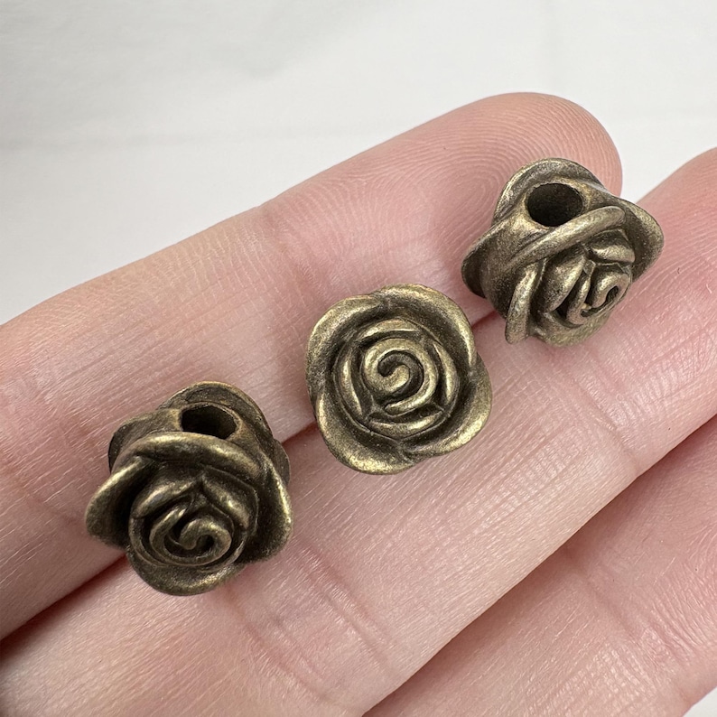 May include: Three bronze-colored rose-shaped beads. Each bead has detailed petals and a hole through the center. The beads are small and appear to be made of metal.