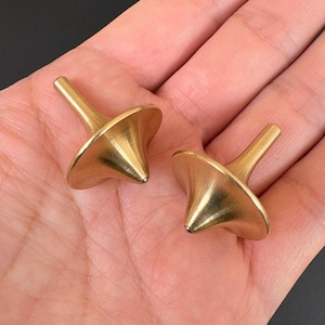 May include: Two brass-colored spinning tops with a classic design. Each top has a conical body and a slender, cylindrical handle. The tops are displayed on a neutral background, highlighting their metallic sheen and simple elegance.