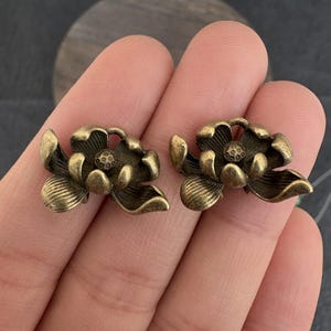 May include: Two antique brass-colored lotus flower-shaped earrings. Each earring features detailed petals and a central seed pod design. The earrings are presented on a neutral background.