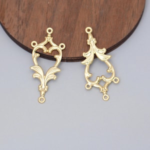 May include: Two gold-coloured filigree charms with ornate, symmetrical designs. Each charm features a central open shape with decorative flourishes and multiple small loops for attachment. The charms are likely for jewellery making or other craft projects.
