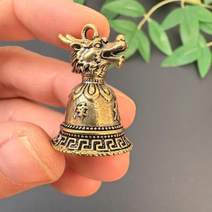 Può includere: Una piccola campana antica in ottone con un design a testa di drago. La campana presenta dettagli intricati, tra cui un volto di drago, motivi decorativi e un anello per appenderla. La campana è tenuta in mano.