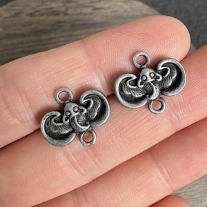 Puede incluir: Dos pequeños dijes de metal en tono plateado con forma de mantarrayas. Cada dije presenta un diseño detallado y en relieve de una criatura acuática con un cuerpo central, aletas anchas y dos bucles circulares en la parte superior e inferior.