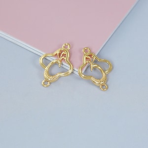 Könnte beinhalten: Zwei goldfarbene Schmuckanhänger mit herzförmigem Design. Jeder Anhänger hat eine strukturierte, durchbrochene Herzform mit einem kleineren Herzdetail oben und einer Schlaufe oben und unten zum Befestigen.