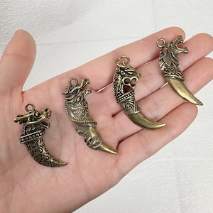 May include: Four antique brass-colored pendants in the shape of dragon teeth. Each pendant features a detailed dragon head design with varying textures and styles. The pendants are suspended from small loops, ready for use in jewelry or crafts.