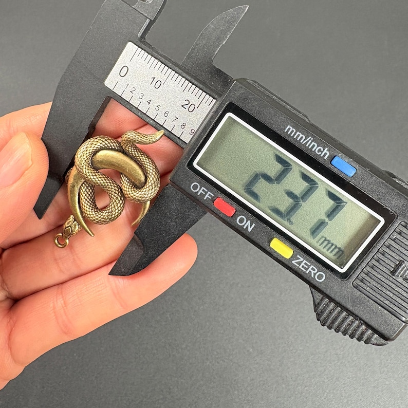 May include: A brass-colored snake-shaped pendant is measured with a digital caliper. The digital display reads 23.7 mm. The pendant is coiled with a detailed scale pattern.
