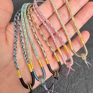Pode incluir: Uma coleção de cinco cordões trançados com várias combinações de cores, incluindo cinza, azul, marrom, rosa e dourado metálico. Cada cordão termina com um laço de cor sólida na base, fixado com um fio dourado enrolado.