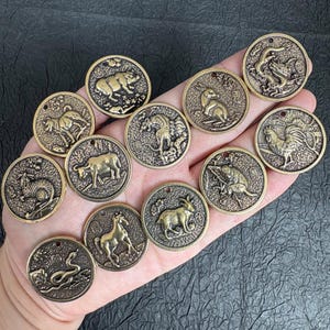 Puede incluir: Una colección de doce dijes redondos de metal en tono bronce, cada uno con un diseño en relieve de un animal diferente del zodiaco chino. Cada dije circular incluye un pequeño orificio en la parte superior para sujetarlo y un fondo texturizado.