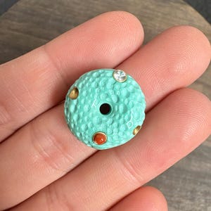 May include: A round, turquoise-colored bead with a textured, cratered surface. The bead features a central hole and is accented with three small, circular insets: one clear, faceted crystal and two smooth, reddish-orange stones set in gold-toned metal.
