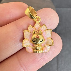 May include: A gold-coloured pendant depicting a stylized cat within a lotus flower. The flower petals are pink with glitter accents. The cat has black eyes and a red heart on its forehead. The pendant has a loop for a chain.