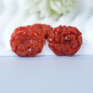 Puede incluir: Tres colgantes de flores de peonía talladas en piedra preciosa roja. Los colgantes son muy detallados, con pétalos visibles y un núcleo floral central. El fondo es blanco suave y azul claro.