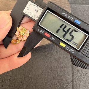 May include: A gold pendant with a pink and white flower design, measured with a digital caliper. The caliper shows 14.5 mm. The pendant has a loop for a chain. The measurement is approximately 0.57 inches.
