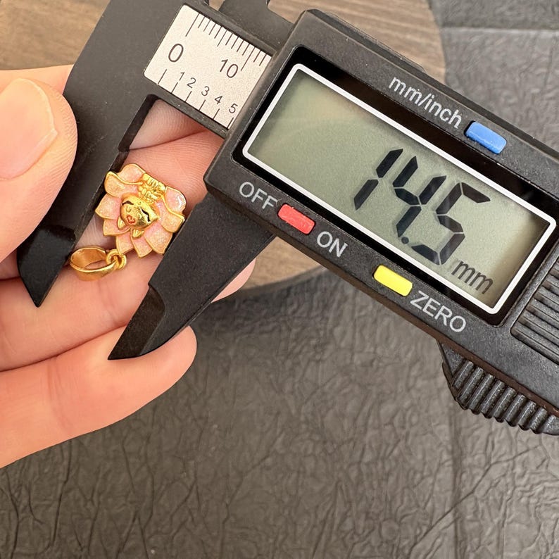 May include: A gold pendant with a pink and white flower design, measured with a digital caliper. The caliper shows 14.5 mm. The pendant has a loop for a chain. The measurement is approximately 0.57 inches.