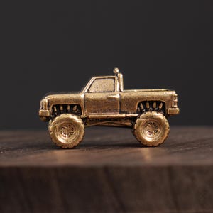 May include: A small, detailed bronze-colored toy truck with large off-road tires. The miniature pickup truck has a cab, a bed, and a small exhaust pipe. The truck is sitting on a wooden surface.