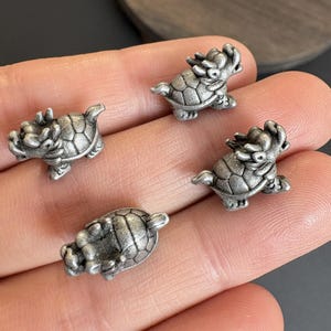以下が含まれることがあります： 4つの小さな銀色のドラゴンのカメの置物。各置物は、詳細な甲羅、ドラゴン風の頭、小さな脚を持っています。置物はニュートラルな背景に表示されています。