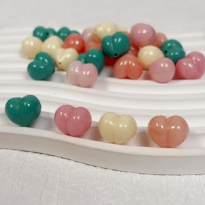 May include: A collection of heart-shaped beads in various colors, including teal, pink, peach, and cream. The beads are arranged on a white, wavy surface, with some scattered in the background. The beads are smooth and glossy.