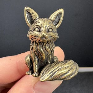 May include: A detailed brass fox figurine with a sitting pose, featuring a textured surface and a bushy tail. The fox has large ears and a detailed face. The figurine is a decorative item.