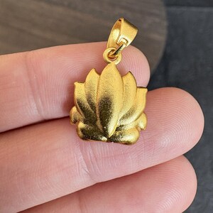 May include: A gold-coloured pendant shaped like a lotus flower. The pendant has a loop at the top for a chain. The lotus flower has detailed petals and a textured surface.