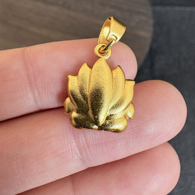 May include: A gold-coloured pendant shaped like a lotus flower. The pendant has a loop at the top for a chain. The lotus flower has detailed petals and a textured surface.