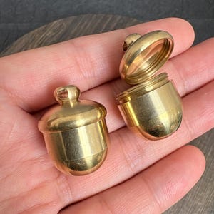 Könnte beinhalten: Zwei kleine, eichelförmige Messingbehälter mit poliertem Goldton-Finish. Ein Behälter ist geschlossen, während der andere geöffnet ist und einen hohlen Innenraum sowie einen Schraubmechanismus zeigt. Jedes Stück hat eine kleine Öse an der Oberseite.