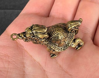 May include: A detailed brass figurine of a mythical creature, a hybrid of a dragon and a turtle. The figure has intricate details, including wings, a dragon head, and a turtle shell with a Yin Yang symbol. The figurine is held in a hand.