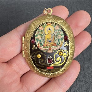 Könnte beinhalten: Ein ovales Medaillon aus goldfarbenem Metall mit einer zentralen runden Einlage einer sitzenden Buddha-Figur. Die dunkelbraune Oberfläche ist mit gelben Wolkenmotiven, kleinen roten Edelsteinakzenten und einem strukturierten Rand verziert.