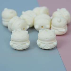 May include: A collection of small, off-white cat figurines. The figurines have a rounded shape with detailed facial features, including closed eyes and a small nose. Each cat has a decorative band around its middle and a star-like design on its belly.