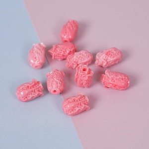 May include: A collection of pink, textured beads scattered on a two-tone pink and blue surface. The beads have a cabbage-like appearance, with detailed ridges and a glossy finish. The beads are likely for jewelry making or crafting.